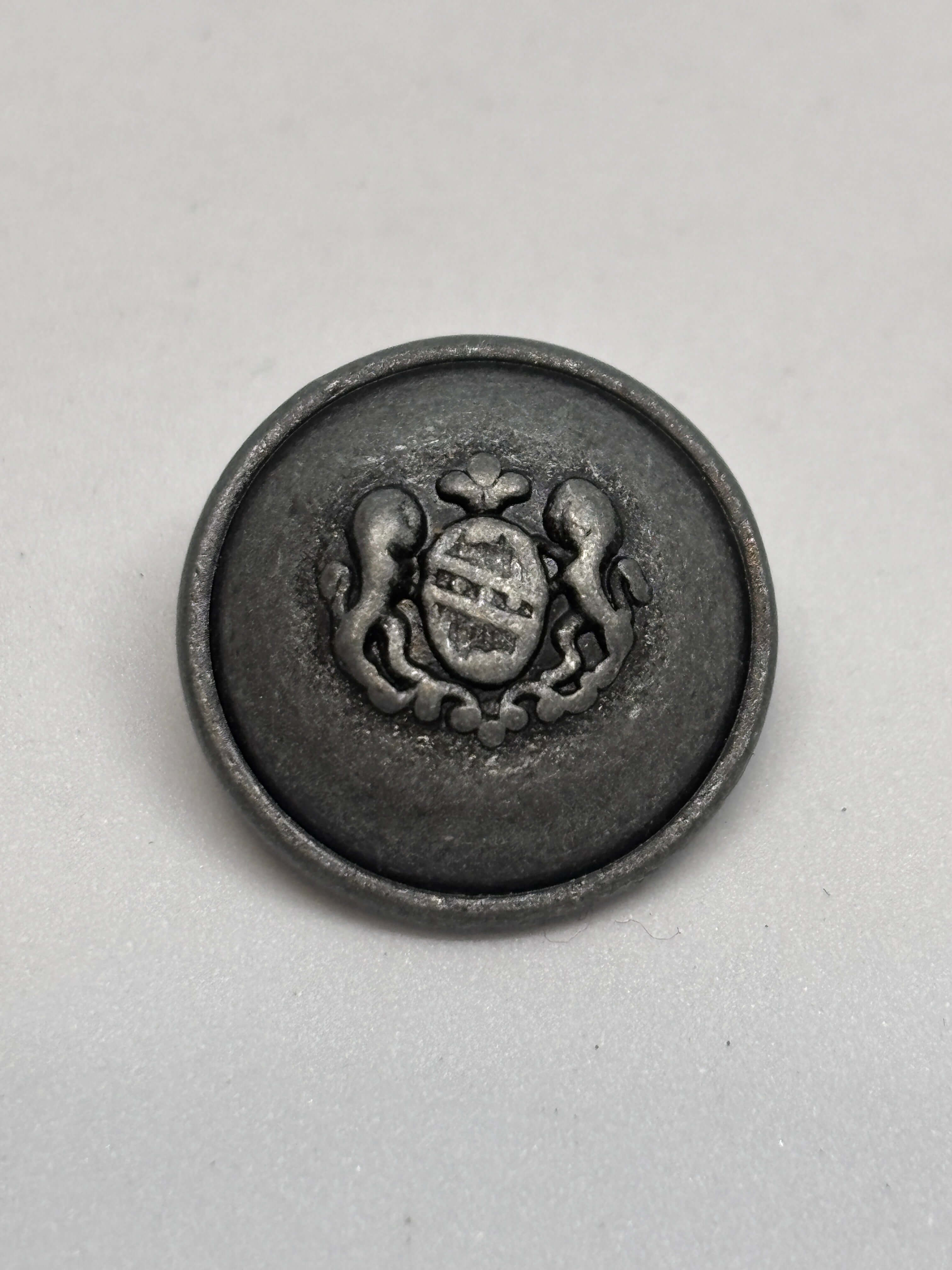 Botón 54549 de vástago en plata antigua con diseño de cresta heráldica, ideal para prendas medianas. button