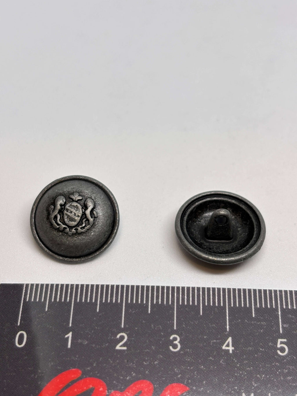 Botón 54549 de vástago en plata antigua con diseño de cresta heráldica, ideal para prendas medianas. button